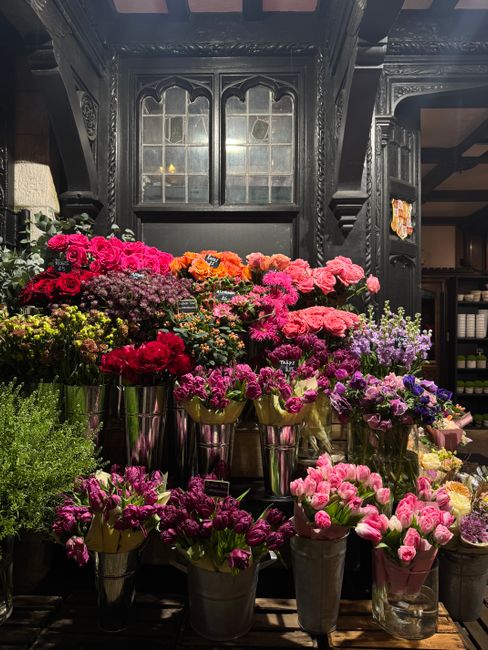 A vibrant selection of fresh flowers arranged in metallic vases and containers, featuring pink, purple, red, orange, and yellow blooms such as carnations, chrysanthemums, and petunias. The flowers are
