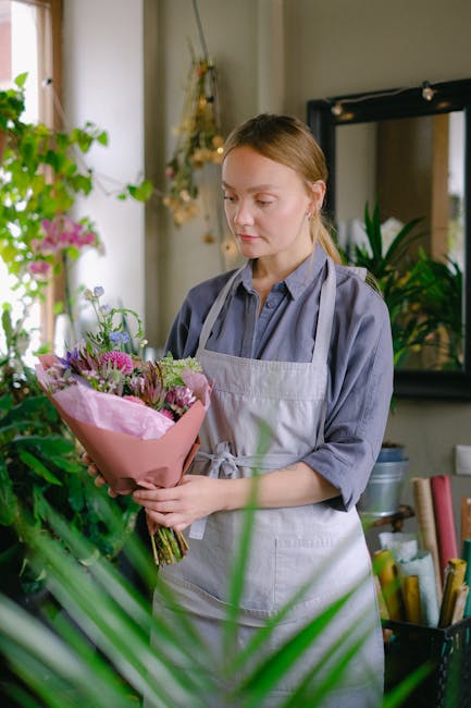 A young woman with light brown hair, wearing a light grey apron over a blue shirt, is holding a bouquet of fresh flowers wrapped in pink paper. The bouquet features a mix of pink, purple, and green bl