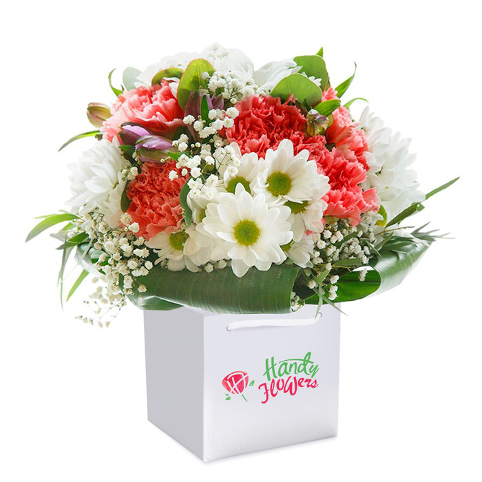 This sophisticated bouquet, arranged with the artistry of a local St Albans florist, takes centre stage within a clean, square white gift bag-its modern lines softly illuminated against a pure white backdrop evocative of gallery space along St Peter's Street. Coral and peach carnations, their abundant frilly petals tenderly layered, form the colourful foundation, sending gentle ripples of warmth through the composition. Scattered among them are pristine white daisies, their distinctive lime-hued centres bright and lively, recalling wildflowers blooming near the River Ver. Wispy tendrils of white baby's breath thread delicately around the larger flowers, imparting a weightless freshness, while just to the side, a handful of tightly closed purple-tipped buds add dimension and a secretive charm. The whole bouquet is encased by a flourish of glossy green foliage; leaves ranging from broad and shining to slim and structured create an elegant natural collar, supporting and highlighting the interplay of colours above. The white bag's robust ribbon handles are tailored for gift giving, suitable for hand delivery to loved ones anywhere from Cottonmill Lane to The Quadrant. Each floral stem has been placed with intention, shaping a full, rounded bouquet that speaks of abundance and thoughtful curation-ideal for birthdays, anniversaries, or as a spontaneous gesture of kindness across St Albans. The distinct branded marking on the bag subtly affirms the artistry and reliability of local floral service, making this arrangement a refined, memorable choice for any significant moment.