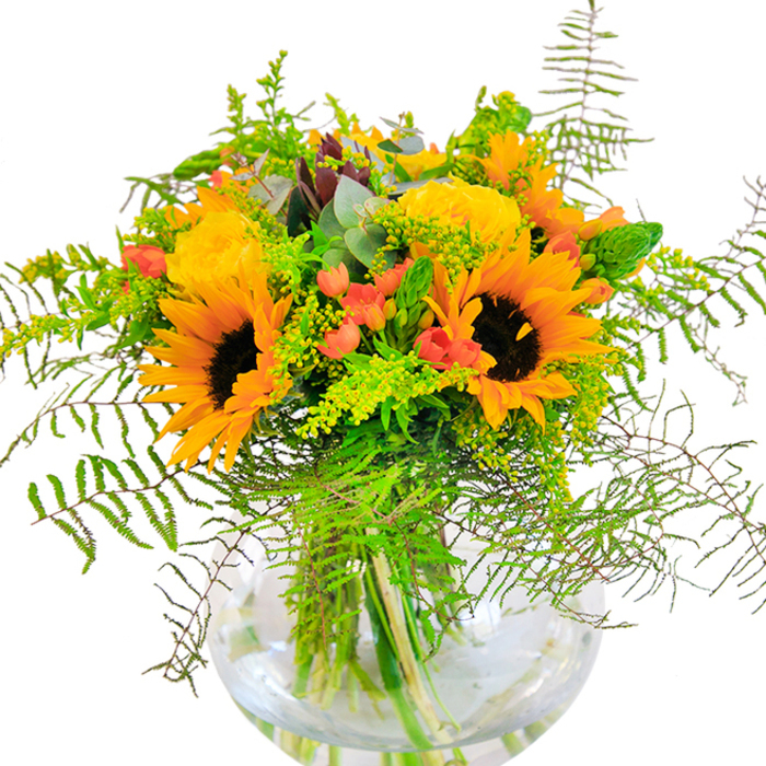 This exuberant bouquet offers a first impression of radiant joy, as vivid yellow sunflowers and garden roses stretch open in sun-drenched layers, their warmth infusing any space with light. The plush, bright petals are companioned by clusters of red Hypericum berries, sparkling among deep greens and airy tendrils-reminiscent of the colorful displays one might find amid St Albans' local flower stalls. Wisps of fern and light filler blossoms extend outward in a sweeping arc, giving the arrangement a lush, garden-fresh feel that calls to mind strolls near the Abbey's green lawns. Eucalyptus leaves add a cool, blue-green contrast, their rounded tips softening the bouquet's energy. Every stem and flourish is anchored within a transparent, rounded glass vase that amplifies the visual brightness and elegant lines of the arrangement. Up close, you'd notice the interplay of textures-the suede-like sunflowers, the tightly spiraled roses, the crisp snap of the verdant greens. The bouquet exudes a gentle, uplifting scent reminiscent of warm fields at the city's edge. Ideal for birthdays, anniversaries, or heartfelt moments of congratulations, this arrangement brings the abundant beauty of St Albans' floral heritage into the heart of every celebration.